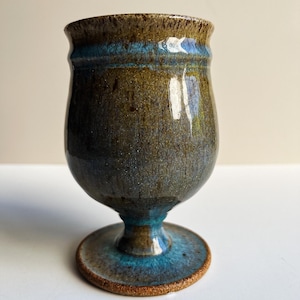May include: A ceramic goblet with a glossy finish, featuring a mottled blend of earthy brown and deep olive green tones. A distinct horizontal band of vibrant turquoise blue encircles the upper rim, while the circular base shows a textured brown edge.