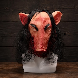 Pode incluir: Uma máscara de cabeça inteira com um rosto de porco rosa realista, pele texturizada, orelhas proeminentes e cabelo sintético preto e ondulado. A máscara possui traços faciais definidos, incluindo aberturas para os olhos e um focinho.