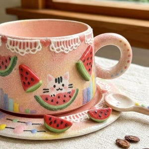Puede incluir: Un juego de taza y plato de cerámica rosa con rodajas de sandía en relieve y una pequeña ilustración de un gato. La taza presenta un patrón de borde similar al encaje blanco, un asa redondeada y una pequeña cuchara de cerámica a juego.