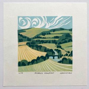 May include: A square linocut print titled Peebles Viewpoint by Katherine Bell, numbered 6/13. The artwork depicts a rolling landscape with yellow fields, dark green trees, a stone bridge over a river, and stylised white clouds in a blue sky.