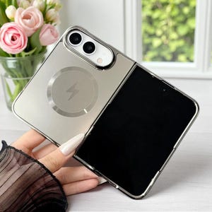 Puede incluir: Un teléfono inteligente plegable con un acabado exterior plateado metálico, un anillo de carga magnético circular y un icono de rayo en el panel posterior. El dispositivo cuenta con un sistema de cámara dual y una pantalla negra rectangular grande.