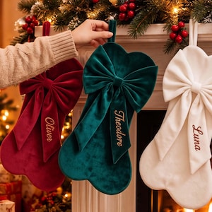 May include: Three velvet Christmas stockings in deep red, dark green, and white, each featuring a large decorative bow and personalized embroidery. The red stocking reads Oliver, the green reads Theodore, and the white reads Luna in gold script.