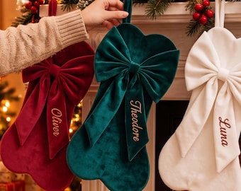 May include: Three velvet Christmas stockings in deep red, dark green, and white, each featuring a large decorative bow and personalized embroidery. The red stocking reads Oliver, the green reads Theodore, and the white reads Luna in gold script.