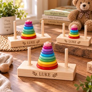 Puede incluir: Tres juguetes de madera para apilar con anillos coloridos en rojo, naranja, amarillo, verde, azul claro, azul oscuro, morado y rosa. Cada base es de madera clara y presenta el nombre LUKE grabado con detalles de hojas en la parte frontal.