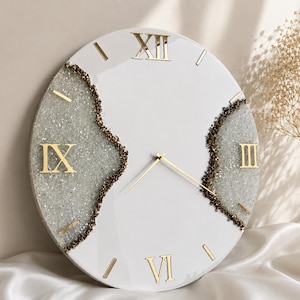 May include: A round wall clock with a smooth white face and gold Roman numerals. Two textured, silver-toned sections with dark brown edges run vertically across the clock face. The clock features thin, gold-colored hour and minute hands.