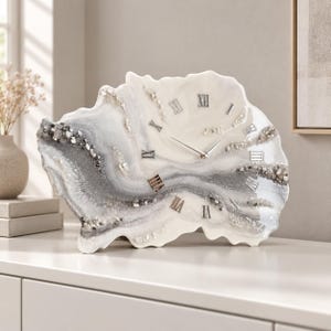 Può includere: Un orologio in resina in stile geode con una forma organica irregolare. La superficie presenta vortici di toni bianchi, grigi e argento con frammenti di vetro e pietra incastonati. Include numeri romani argentati e sottili lancette d'argento.