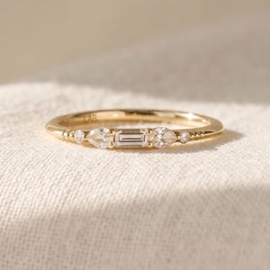 May include: A gold ring featuring a central baguette-cut stone flanked by two pear-cut stones and two small round stones. The band is thin and polished, with the text 14K LAB engraved on the inner surface of the metal band.