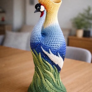 May include: A crochet vase shaped like a crowned crane bird. The base features green textured grass patterns, transitioning into a blue sky with white clouds. The neck of the vase is yellow, topped with a golden fan-like crest and a crane head.