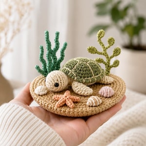 May include: A crocheted sea turtle figurine resting on a circular tan base. The turtle has a green shell and cream-colored body. The base features a small orange starfish, two tiny seashells, and two upright green crocheted aquatic plant structures.