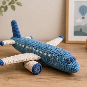 Puede incluir: Un avión de juguete hecho a mano en ganchillo con fuselaje azul y alas blancas. La aeronave tiene puntas de ala azules, detalles de motor azules y una ventana de cabina azul oscuro. El cuerpo está texturizado con puntos de hilo.