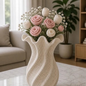 May include: A cream-colored crocheted vase with a spiraling, ruffled rim design. The vase holds a bouquet of handcrafted crocheted flowers, including pink roses, white tulips, and small white filler flowers with green crocheted stems and leaves.