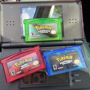 May include: A black Nintendo DS handheld console with three Game Boy Advance video game cartridges resting on its surface. The cartridges are translucent green Pokémon Emerald, translucent red Pokémon Ruby, and translucent blue Pokémon Sapphire.