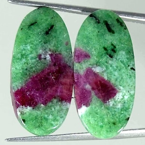 Puede incluir: Dos cabujones de forma ovalada de piedra preciosa rubí zoisita. Cada piedra presenta un color base verde vibrante con parches irregulares distintos de inclusiones de rubí rojo magenta intenso y pequeñas motas minerales negras.