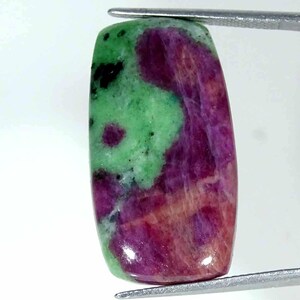 Può includere: Una pietra preziosa lucidata di forma rettangolare che presenta un mix vibrante di inclusioni minerali color magenta intenso e verde brillante. La pietra mostra una superficie liscia e lucida con zone di colore distinte e piccole macchie nere.