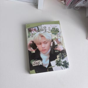 May include: A rectangular photocard featuring a portrait of a person with blonde hair wearing a white turtleneck and black jacket, holding black headphones. The design includes green clover illustrations, the text "Jay", "04", "20", and Chinese characters.