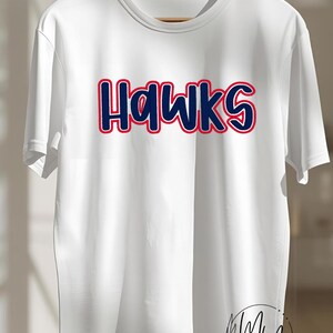 May include: A white short-sleeved t-shirt featuring the word HAWKS printed in dark blue letters with a red outline across the chest. A circular logo with the letters MM and the text Mommie's Monogramming & Co. is visible at the bottom right.