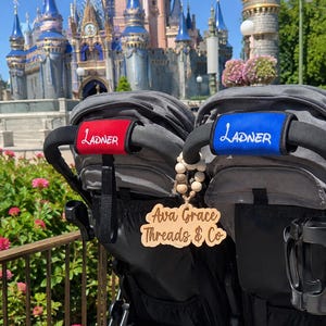 May include: A double stroller featuring two black handles with personalized covers in red and blue, each embroidered with the name Ladner in white. A wooden tag hanging from the handle displays the text Ava Grace Threads & Co in script.
