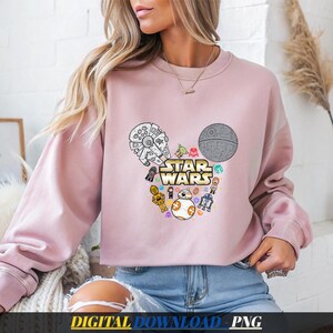 Könnte beinhalten: Ein hellrosa Langarm-Sweatshirt mit einer Grafik des Star Wars-Logos, umgeben von verschiedenen Charakteren und Raumschiffen in Form von Mickey-Mouse-Ohren, darunter der Millennium Falke, der Todesstern, BB-8 und C-3PO.