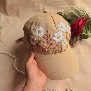 May include: A tan baseball cap featuring detailed embroidery of white daisies with yellow centers and small pink floral accents. The embroidery is positioned on the front panels of the cap, showcasing a botanical design with green stems.