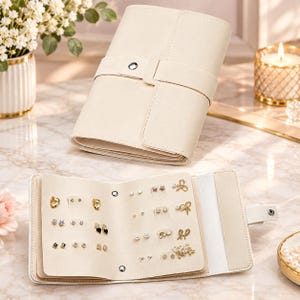 May include: A cream-colored, foldable jewelry organizer case made of smooth material with a snap-button closure. The interior features multiple pages with pre-cut holes for storing various pairs of stud earrings and small dangling earrings.