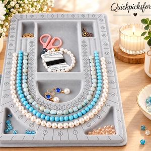 May include: A gray flocked jewelry beading board with U-shaped channels, numbered markings from 0 to 22, and recessed compartments. It contains strands of white and blue beads, a pair of pink scissors, a spool of silk thread, and gold earrings.