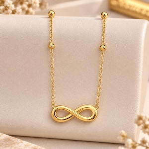 May include: A gold-toned necklace featuring a central infinity symbol pendant. The chain is composed of delicate oval links and includes two small, smooth gold-toned beads positioned symmetrically on each side of the chain above the pendant.