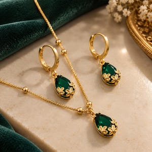 Peut inclure: Un ensemble de bijoux doré composé d'un collier et d'une paire de boucles d'oreilles créoles. Chaque pièce présente une pierre précieuse vert foncé en forme de goutte, sertie dans du métal doré orné de motifs floraux et de petites pierres.