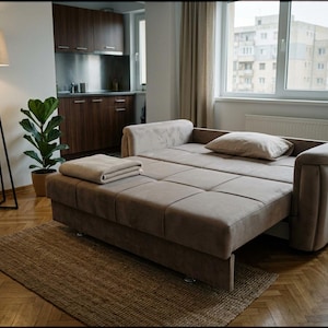 May include: A beige upholstered sofa bed in a partially extended position, featuring a tufted seat surface, rounded armrests, and a matching rectangular pillow. The sofa is covered in a soft fabric and includes a folded light-colored throw blanket.