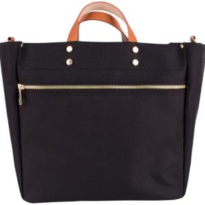 May include: Black tote bag with a gold zipper and brown leather handles. The bag has a rectangular shape with a structured design. Gold rivets secure the handles to the bag. The bag is made of a durable material.