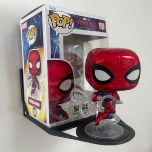 Puede incluir: Una figura cabezona Funko Pop de Spider-Man, número 1590, con un traje rojo y azul metálico y ojos blancos. La figura está colocada sobre una base circular transparente, junto a su caja de cartón original con la marca Marvel.