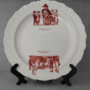 May include: A white ceramic plate with a scalloped edge featuring two red transferware illustrations. The top scene shows a figure with children and the text One a penny, two a penny, Hot Cross Buns. The bottom scene shows figures with fruit.