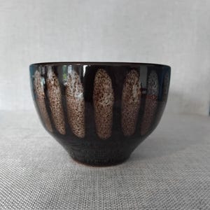 May include: A dark brown ceramic bowl featuring a series of vertical, elongated oval patterns in a lighter, speckled beige tone around the exterior. The bowl has a smooth, glossy finish and a rounded shape that tapers towards a narrow base.