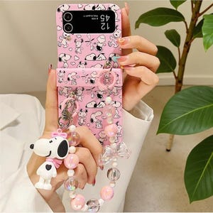 May include: A pink foldable smartphone case featuring a repeating pattern of Snoopy characters. The case includes a matching strap adorned with translucent pink and clear beads, a small clear bow, and a dangling Snoopy figurine charm.