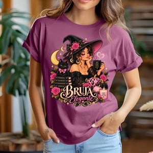 May include: A mauve t-shirt featuring a graphic print of a person wearing a witch hat with a pink rose, a black cat, a stack of books, pumpkins, and a candle. The text reads Bruja Elegante, with smaller text on the mug and candle.