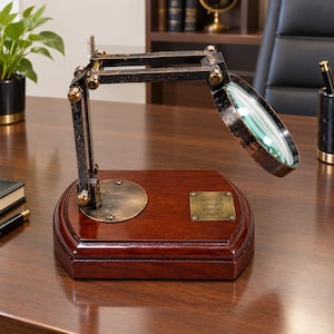 May include: A magnifying glass mounted on an adjustable, distressed black metal arm with brass-toned hardware. The base is a polished, dark wood platform featuring a rectangular brass plaque engraved with the text: WATKINS & SON OPTICIANS LONDON 1925.