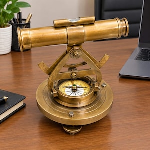 Puede incluir: Un instrumento de topografía náutica de latón que presenta un telescopio horizontal montado sobre una base de brújula circular. El dispositivo incluye un nivel de burbuja integrado sobre el telescopio, un dial giratorio con marcas de grados y una brújula magnética central.