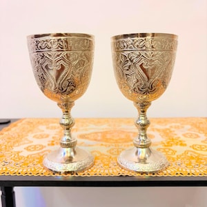Può includere: Due calici in metallo color argento con intricati motivi floreali e a foglia in rilievo sulle coppe e sulle basi. Ogni calice presenta uno stelo sottile e decorativo con un nodo centrale arrotondato e un piede circolare ampio e stabile.