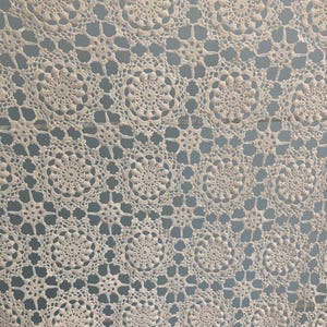 May include: An off-white crochet textile featuring a repeating pattern of circular floral motifs connected by smaller star-shaped elements. The intricate needlework creates a delicate, openwork design with a consistent texture throughout the fabric.