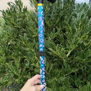 May include: A walking stick featuring a black T-shaped handle, a gold-coloured collar, and a metallic blue shaft decorated with a repeating pink and light blue leopard print pattern. The shaft includes a black adjustable locking mechanism.