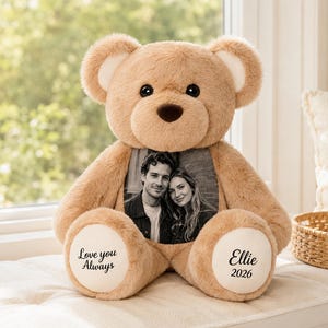 May include: A tan plush teddy bear featuring a black and white photograph of a couple on its chest. The bear has white paw pads with the text "Love you Always" on the left foot and "Ellie 2026" on the right foot, with dark eyes and a nose.