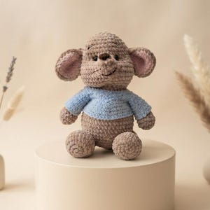 Puede incluir: Un peluche de ganchillo con forma de ratón hecho de hilo marrón claro, con grandes orejas redondeadas con detalles interiores en rosa y una nariz bordada en marrón oscuro. El ratón lleva un suéter de ganchillo azul claro que cubre el torso.