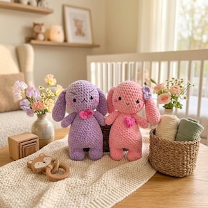May include: Two crocheted plush bunnies, one purple and one pink, each featuring a small heart-shaped accent on the chest. The purple bunny has a stern expression, while the pink bunny has a smiling face and a small purple bow on its ear.