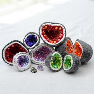 May include: A collection of crocheted geode-inspired objects featuring grey yarn exteriors with white trim and interiors filled with vibrant, multi-colored glass beads in shades of red, purple, orange, and green, alongside two small natural crystals.