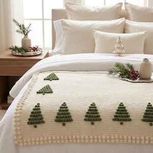 May include: A cream-colored knit throw blanket featuring a repeating pattern of dark green textured pine trees and a border of small raised bobble stitches. The blanket is draped over a bed with white linens and neutral-toned pillows.