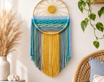 May include: A circular macrame wall hanging featuring a central yellow sun design with radiating cords. The lower section displays horizontal rows of teal, light blue, and sage green knots, finished with long, flowing fringe in yellow, teal, and gray.