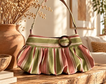 May include: A shoulder bag featuring a pleated design with vertical stripes in shades of sage green, dusty pink, and white. The bag includes a single fabric strap and a prominent dark brown circular ring accent attached to the front panel.