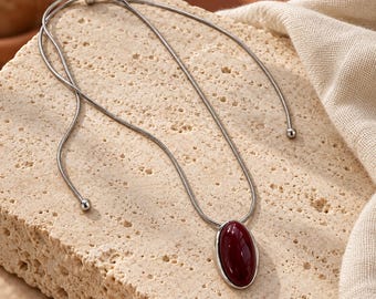 May include: A silver-toned necklace featuring a smooth, oval-shaped deep red cabochon pendant. The chain is a flexible snake-style cord with an adjustable sliding mechanism and small spherical end caps, creating a sleek and polished appearance.