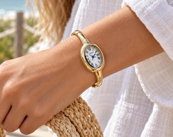 May include: A gold-tone bangle watch featuring an oval-shaped case with a white dial, black Roman numeral hour markers, and blue hands. The watch face displays the brand name NAIDU in black text. The band is a solid, smooth gold-tone metal.