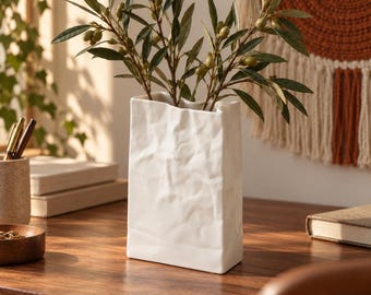 Puede incluir: Un jarrón de cerámica blanca con forma de bolsa de papel arrugada, que presenta una base rectangular y una superficie texturizada y con pliegues. El jarrón contiene varias ramas de olivo verdes con pequeñas aceitunas redondas de color verde claro.
