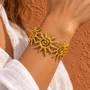 Peut inclure: Un bracelet manchette doré orné d'un motif répétitif de soleils. Chaque soleil présente une spirale centrale et des rayons ondulés. Le métal possède une finition texturée et légèrement irrégulière, formant une bande continue de soleils reliés.